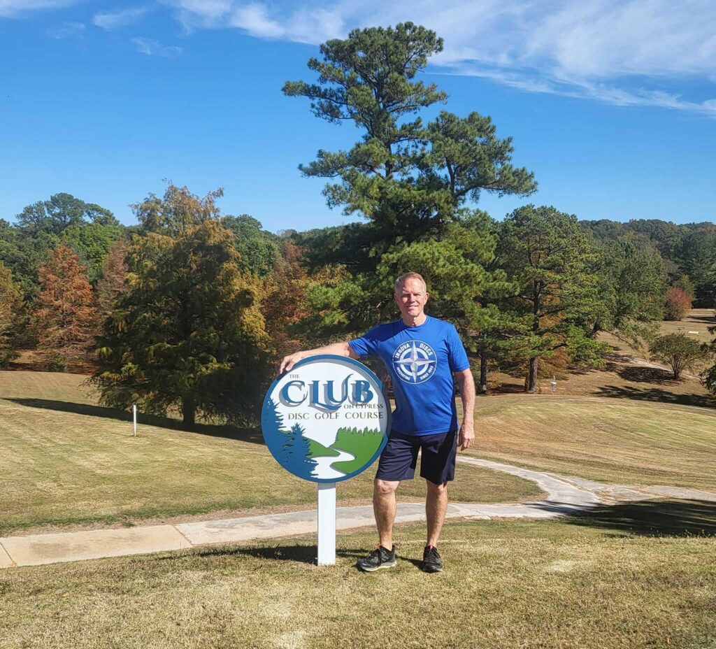 Lavone Wolfe stands on a grassy area next to a Club Disc Golf Course sign. Tall trees and a clear blue sky are in the background.