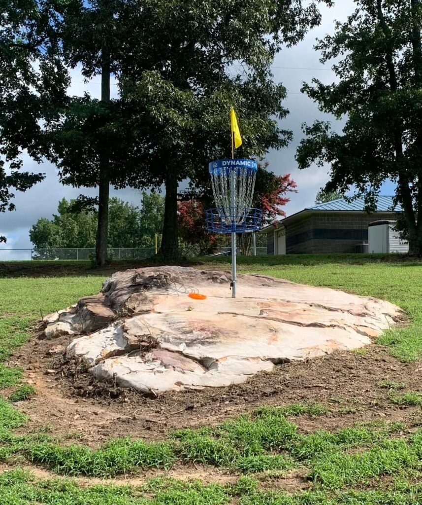 A disc golf course scene with a distinctive rock located at the center, surrounded by grassy terrain and trees.